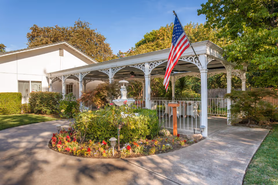Outdoor garden area at Roses & Ivy Assisted Living Facility for the Elderly featuring a white gazebo with decorative trim, an American flag, a white statue fountain surrounded by flowers and greenery, and a paved walkway.
