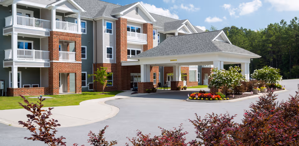 Exterior view of a senior living facility building with a covered entrance, balconies, brick and siding facade, landscaped flower beds, and a paved driveway surrounded by greenery.