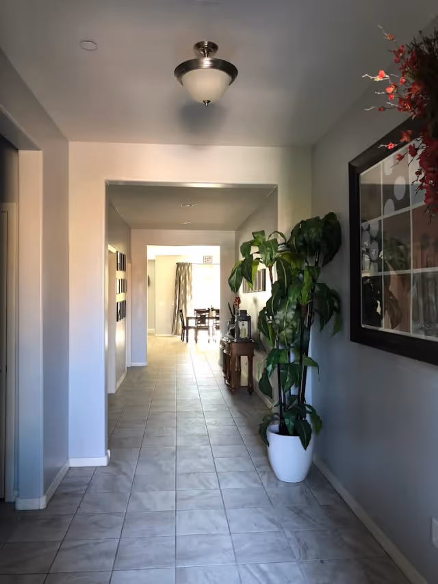 Long tiled hallway inside an assisted living facility with a potted plant, wall art, and a sunlit dining area visible at the end.