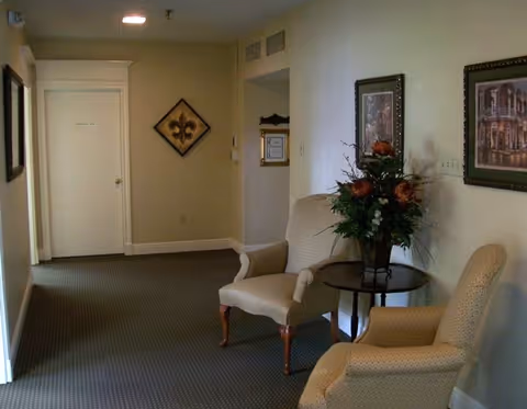 A small seating area in a hallway with two beige upholstered armchairs and a round wooden table between them holding a floral arrangement. The walls are light-colored and decorated with framed artwork. A closed white door is visible at the end of the hallway.