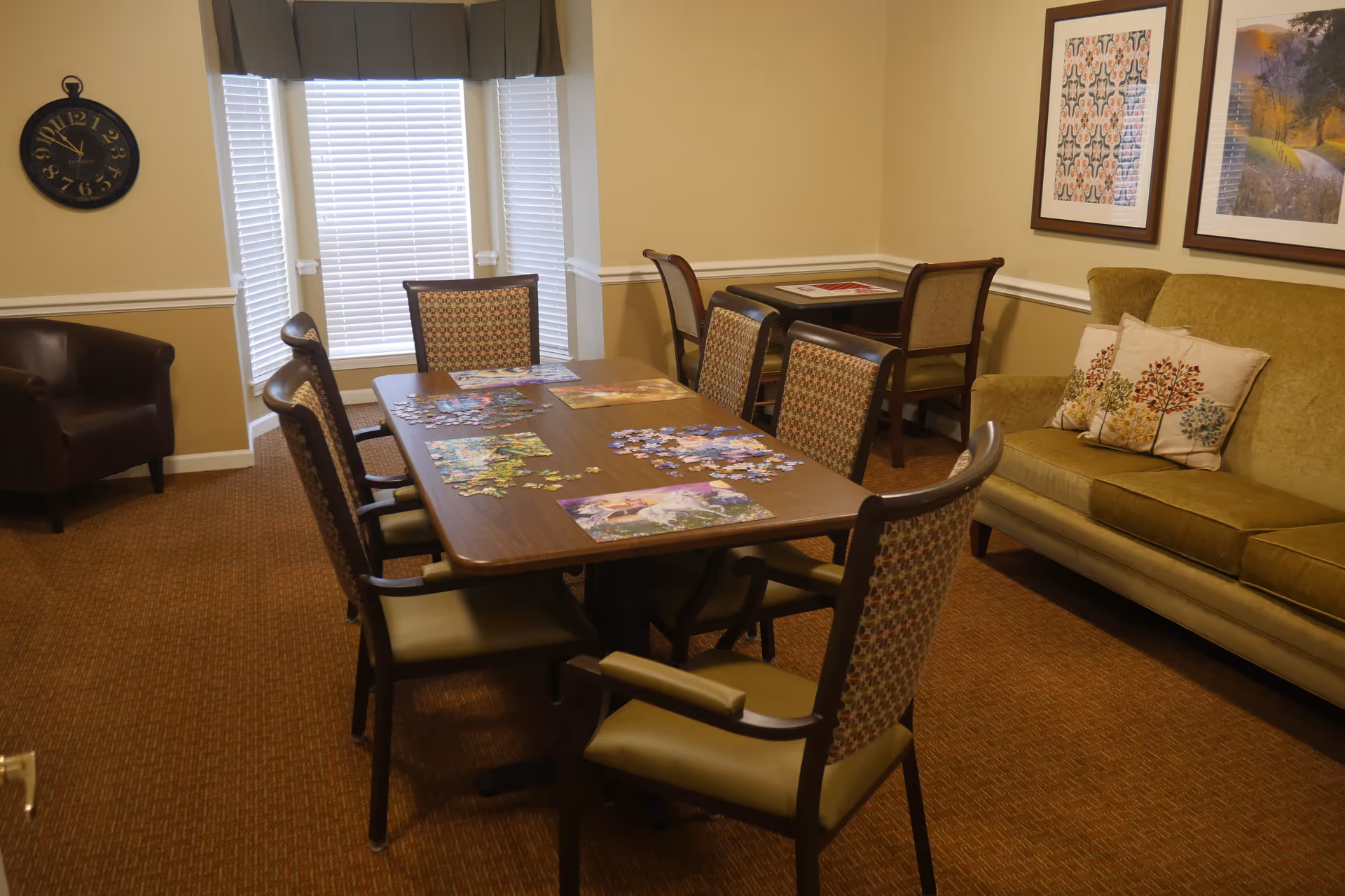 A cozy room with a rectangular wooden table surrounded by six cushioned chairs. On the table, there are several partially completed jigsaw puzzles. To the right, there is a green sofa with three decorative pillows featuring tree designs. The walls are beige with white trim, and two framed pictures hang above the sofa. A brown leather armchair is positioned near a window with white blinds and a dark valance. A wall clock is mounted on the left wall.