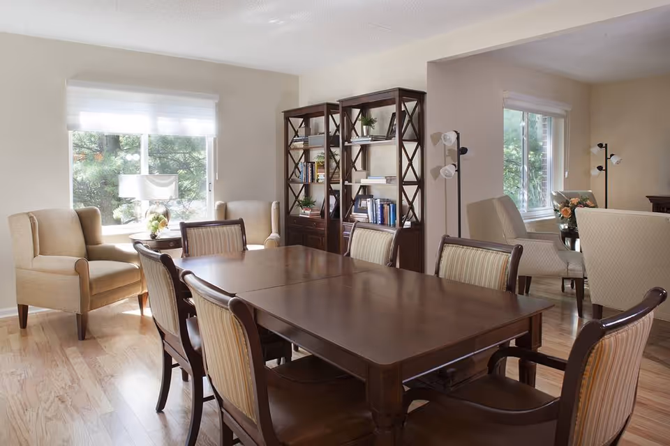 A well-lit dining room with a large wooden dining table surrounded by eight upholstered chairs. In the background, there are two beige armchairs near a window with a small round table and a lamp between them. Two tall wooden bookshelves filled with books and decorative items are against the wall. The room has hardwood flooring and light-colored walls with two windows letting in natural light.