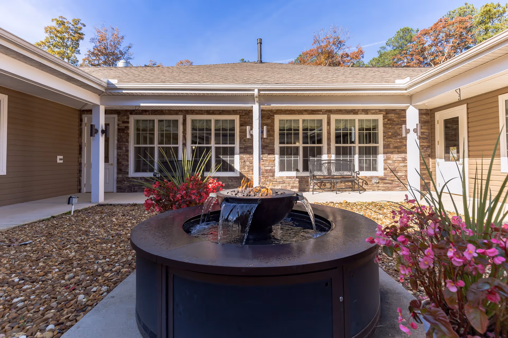 Outdoor courtyard area at Discovery Commons At Wildewood featuring a circular water fountain with fire element in the center, surrounded by landscaping with rocks and flowering plants. The courtyard is enclosed by a building with large windows and a bench visible against the stone and siding exterior walls under a clear blue sky.