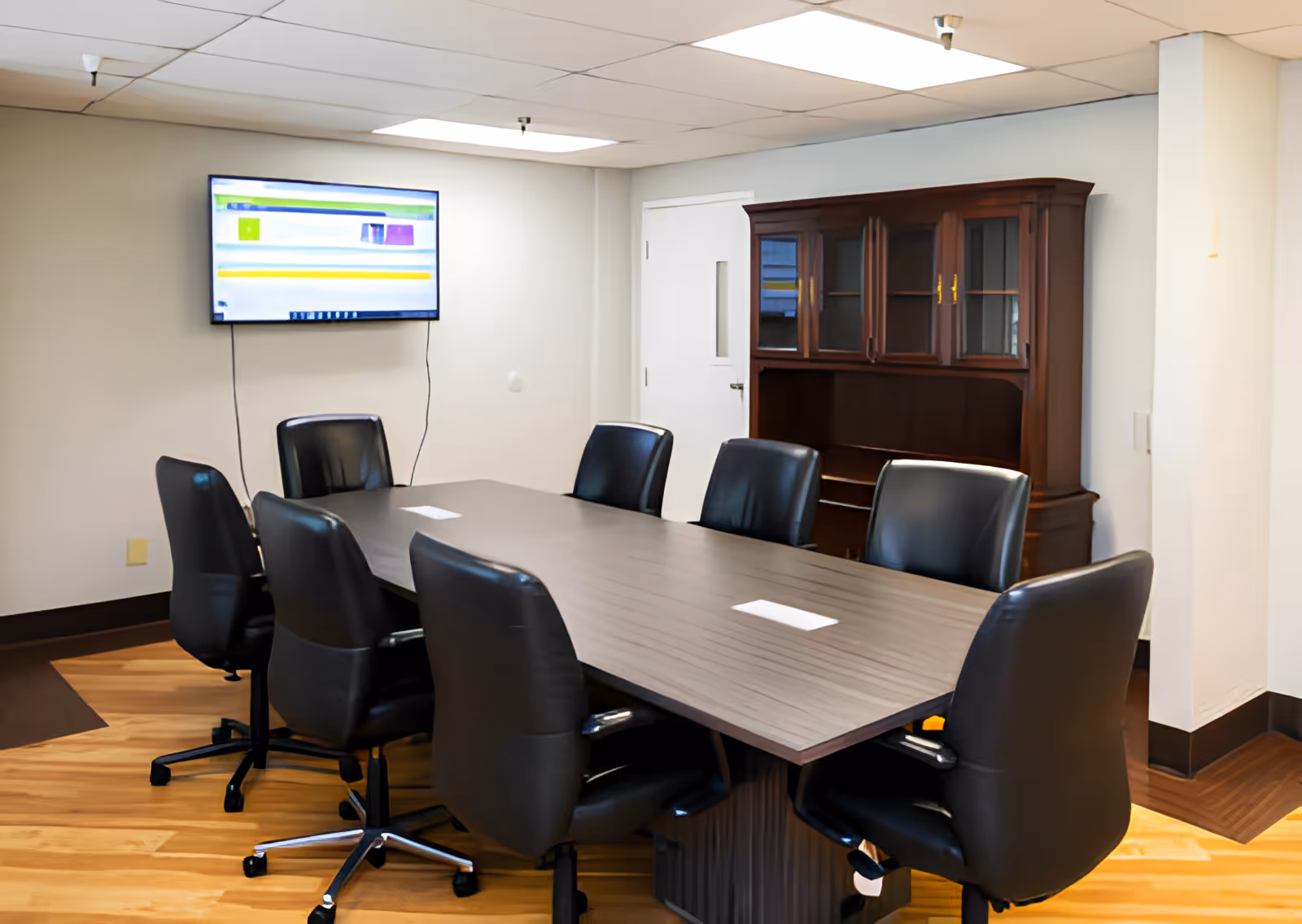 A conference room with a long rectangular table surrounded by eight black office chairs on wheels. A flat-screen TV is mounted on the wall, and a wooden cabinet with glass doors is positioned against the wall. The room has light-colored walls and a wood-patterned floor.