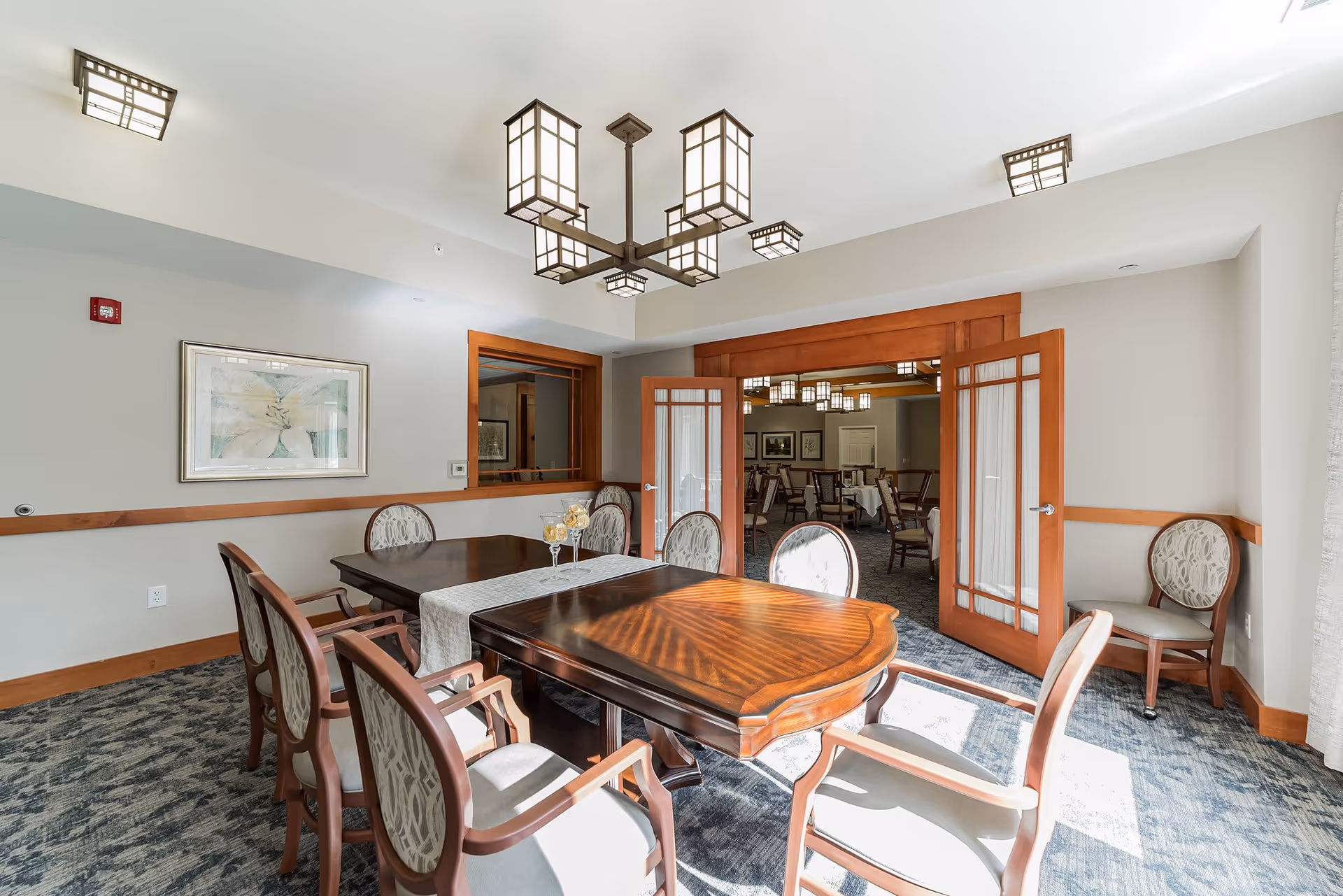 A bright dining room with a wooden table surrounded by six upholstered chairs. The room features a modern chandelier with square lantern-style lights, a framed artwork on the wall, and double wooden doors with glass panels leading to another dining area with more tables and chairs. The floor is carpeted and sunlight streams in through a window on the right.