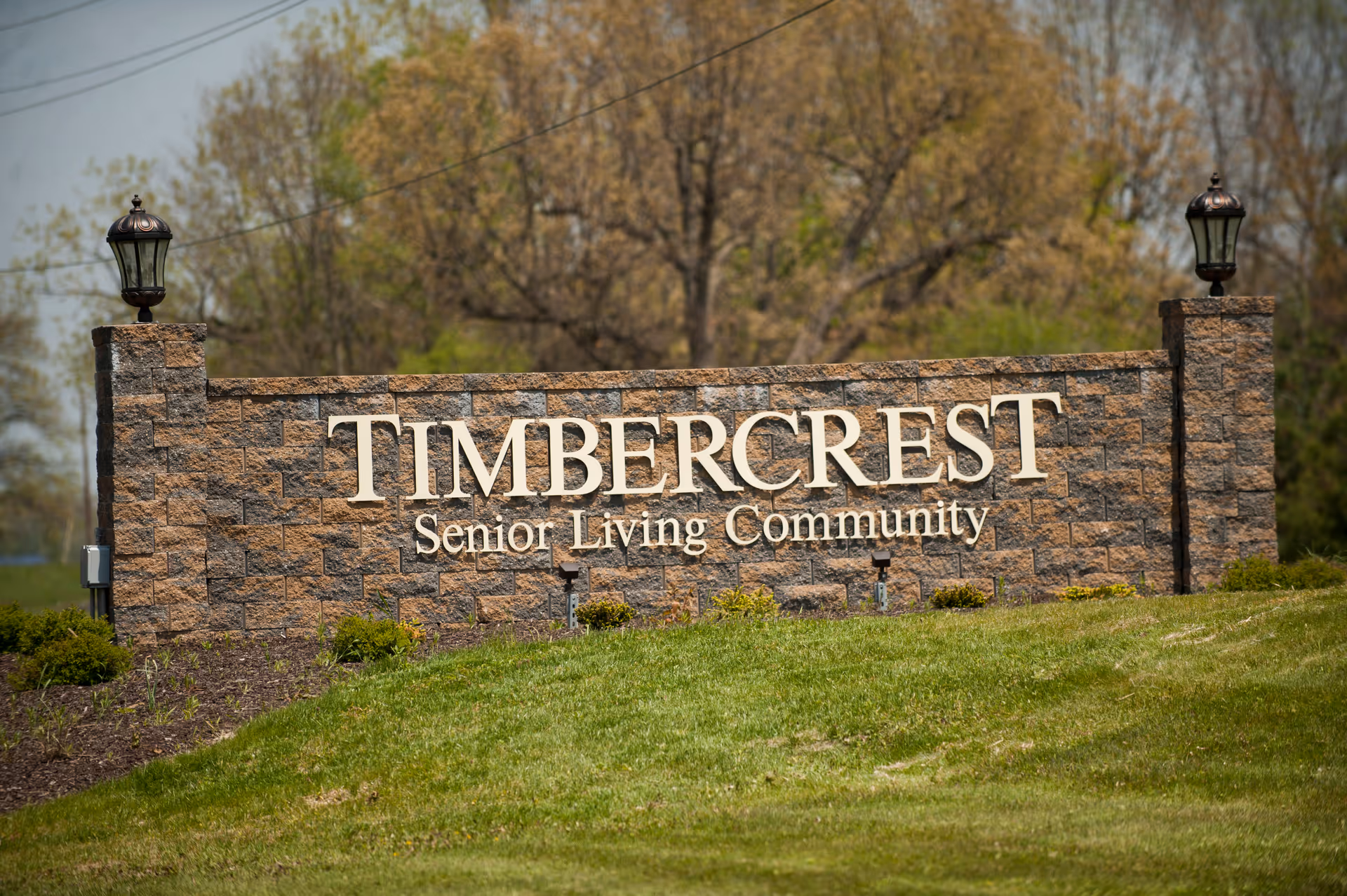A large stone sign with two lanterns on top of pillars on each side, displaying the text 'TIMBERCREST Senior Living Community' surrounded by grass and trees in the background.