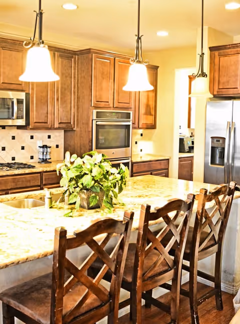 A bright kitchen with wooden cabinets, a granite countertop island with a green leafy plant centerpiece, three wooden bar stools, stainless steel appliances including a refrigerator, oven, and microwave, and three hanging pendant lights above the island.