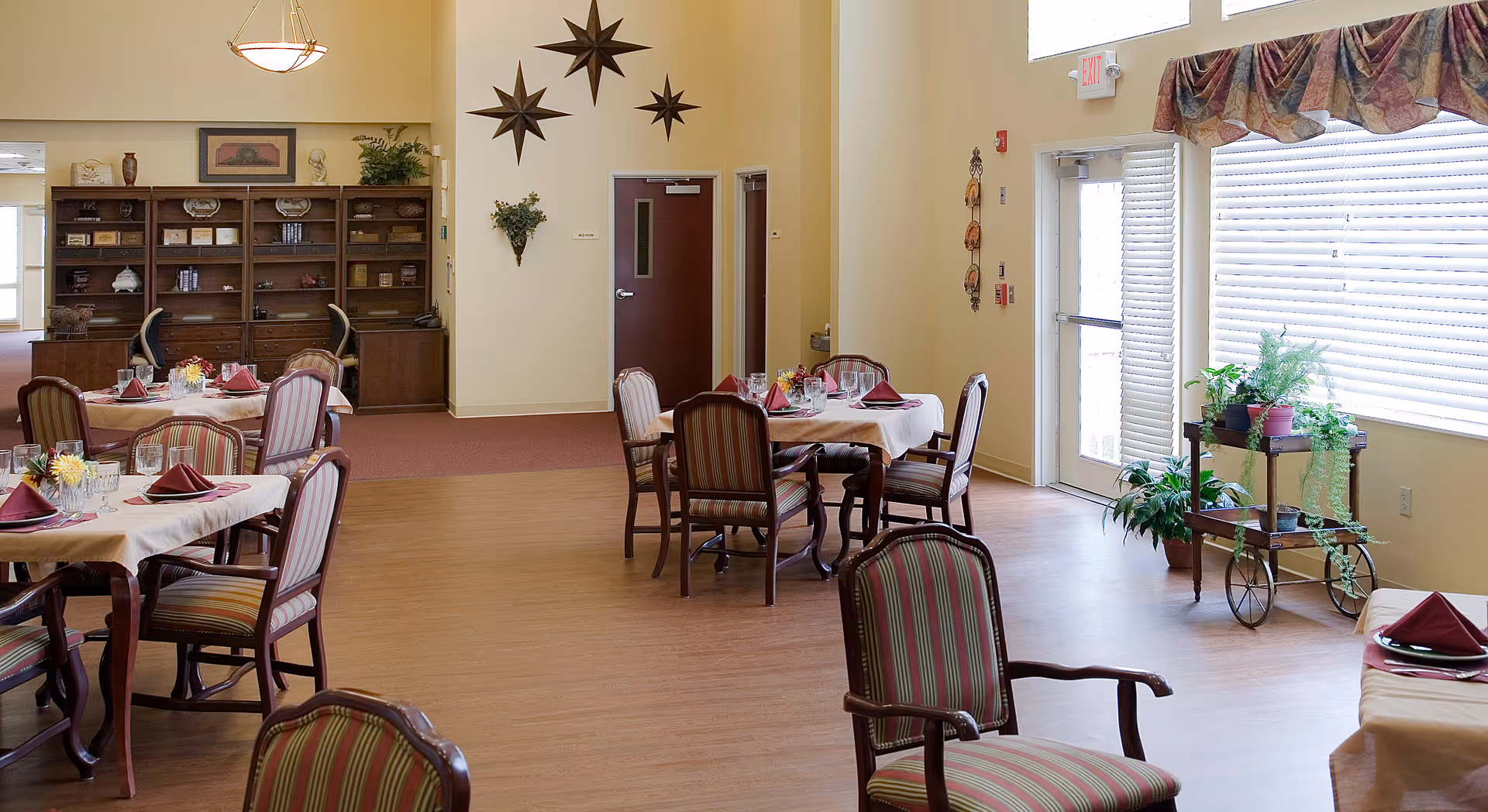 A bright dining room with several tables covered in white tablecloths and set with burgundy napkins, glasses, and plates. The room features striped upholstered chairs, a wooden floor, a large window with blinds and a valance, and a plant stand with various potted plants. On the far wall, there are decorative star-shaped wall hangings and a wooden bookshelf with various items.