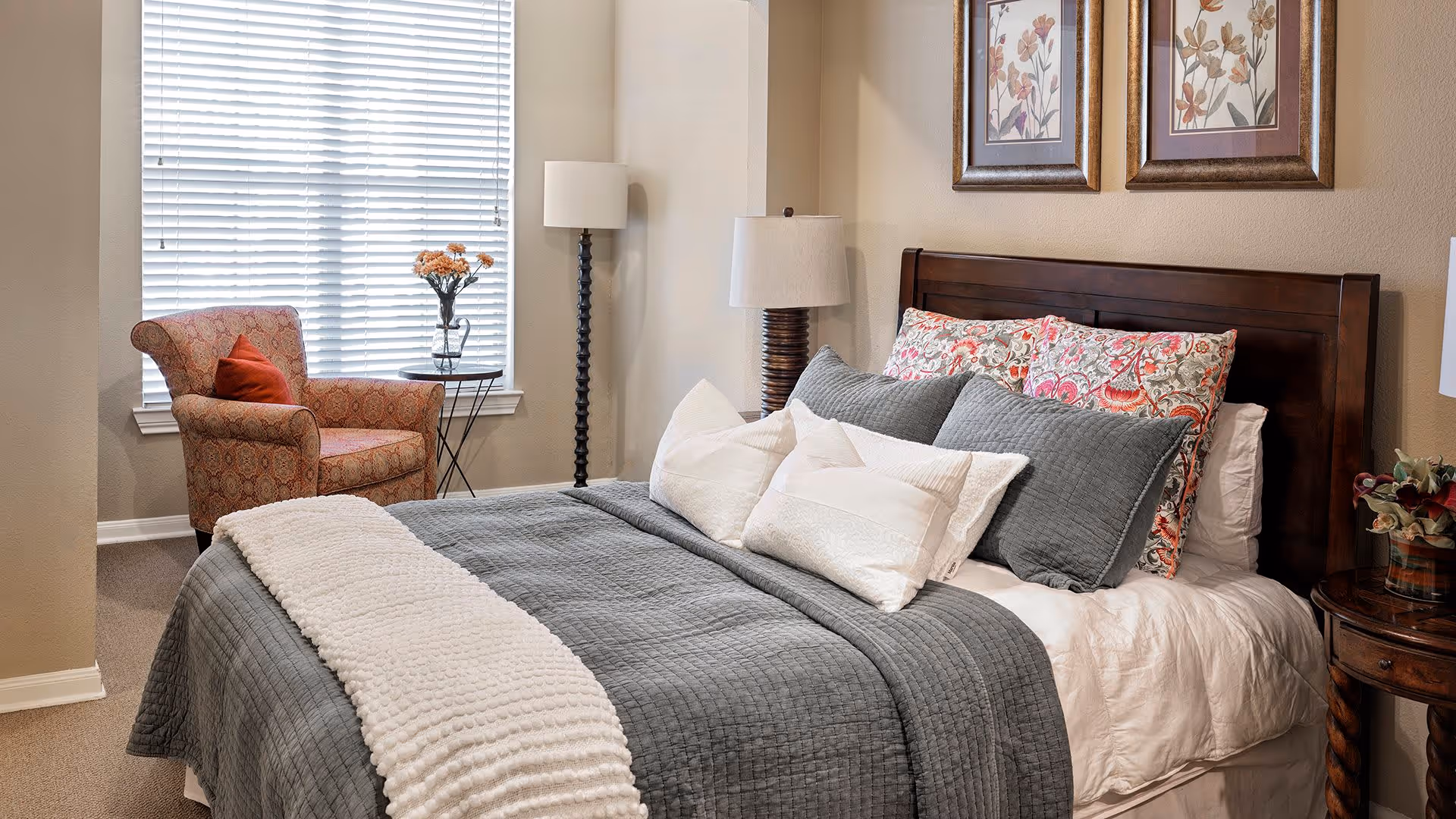 A cozy bedroom featuring a neatly made bed with gray and white bedding, floral-patterned pillows, and a dark wooden headboard. To the left of the bed is a window with white blinds, a patterned armchair with a red cushion, and a small round side table holding a vase with flowers. There are two framed floral artworks on the beige wall above the bed, and a wooden nightstand with a lamp and a small plant on the right side of the bed.