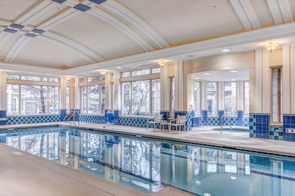 Indoor swimming pool area with large windows allowing natural light to fill the space. The pool is surrounded by blue and white tiled walls and has a pool lift chair for accessibility. There are several chairs and a small table near the pool, and a hot tub area is visible in the background.