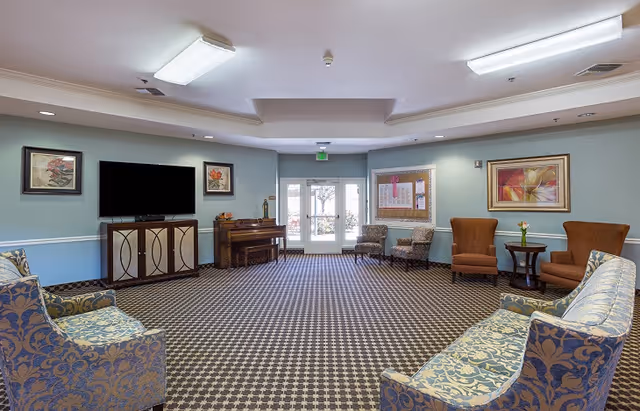 A spacious common area in a senior living facility with patterned carpet, multiple upholstered chairs and sofas arranged around the room, a piano, a flat-screen TV mounted on a cabinet, framed artwork on the walls, and double glass doors leading outside.