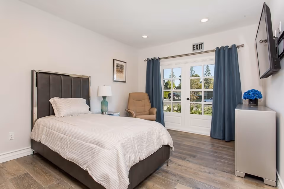 A bright bedroom with a single bed featuring a dark upholstered headboard and light striped bedding. Next to the bed is a nightstand with a greenish-blue lamp. A beige armchair is placed near a set of glass double doors with blue curtains, and an exit sign is mounted above the doors. A flat-screen TV is mounted on the wall opposite the bed, above a small cabinet with a blue flower arrangement. The room has wood flooring and white walls.