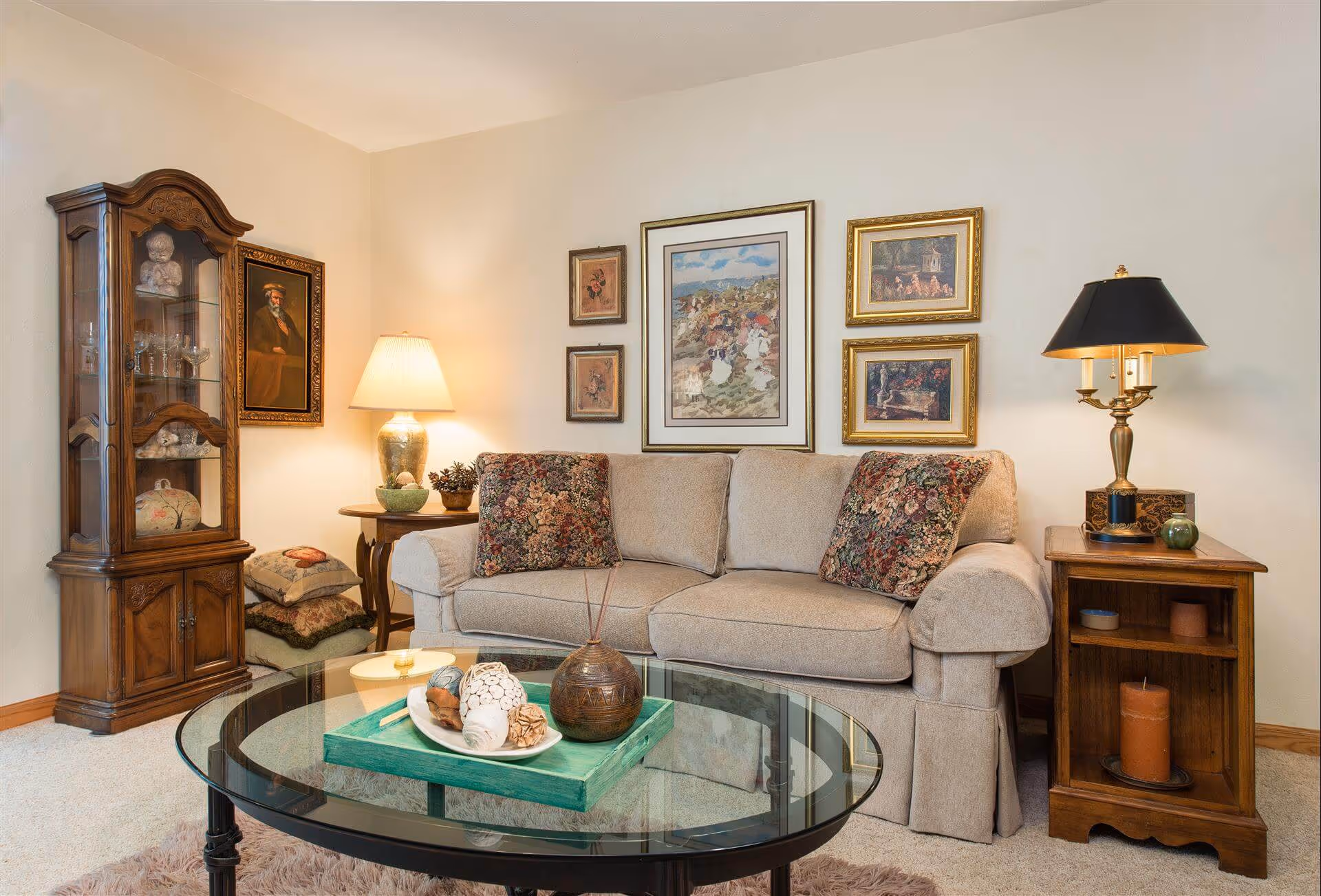 A cozy living room with a beige sofa adorned with two floral patterned pillows. Behind the sofa, there are five framed paintings on the wall. To the left of the sofa is a wooden display cabinet with glass doors containing decorative items, and a side table with a lamp and some cushions stacked underneath. To the right of the sofa is a wooden side table with a black and gold lamp and some small decorative objects. In front of the sofa is a glass coffee table with a green tray holding decorative items.