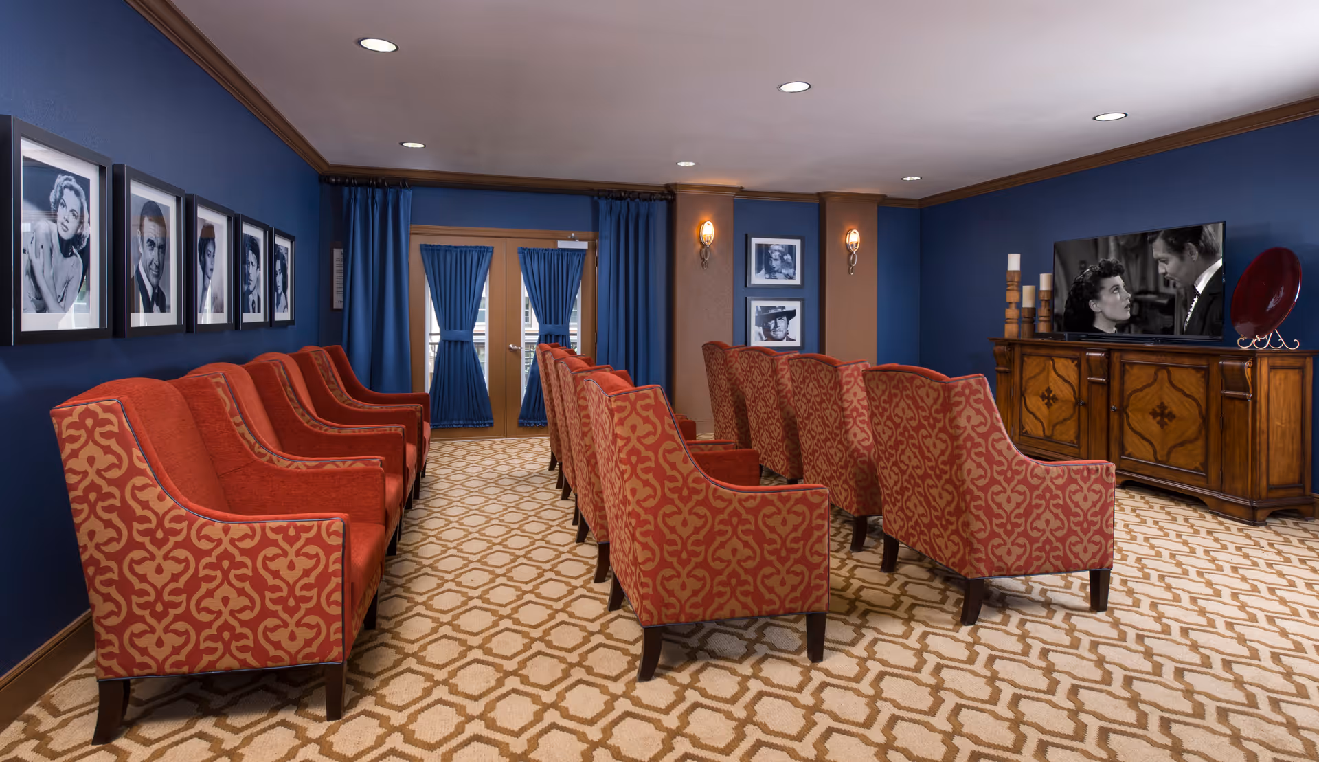 A cozy media room with two rows of red patterned armchairs facing a large flat-screen TV displaying a black and white movie scene. The room has blue walls adorned with black and white portraits of classic film stars, blue curtains on a glass door, and a patterned beige carpet. A wooden cabinet beneath the TV holds decorative candles and a red plate.