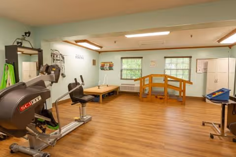 A rehabilitation therapy room with exercise equipment including a stationary bike, a therapy table, a wooden balance bridge, and various therapy tools on the walls. The room has light green walls, two windows with blinds, and wood flooring.