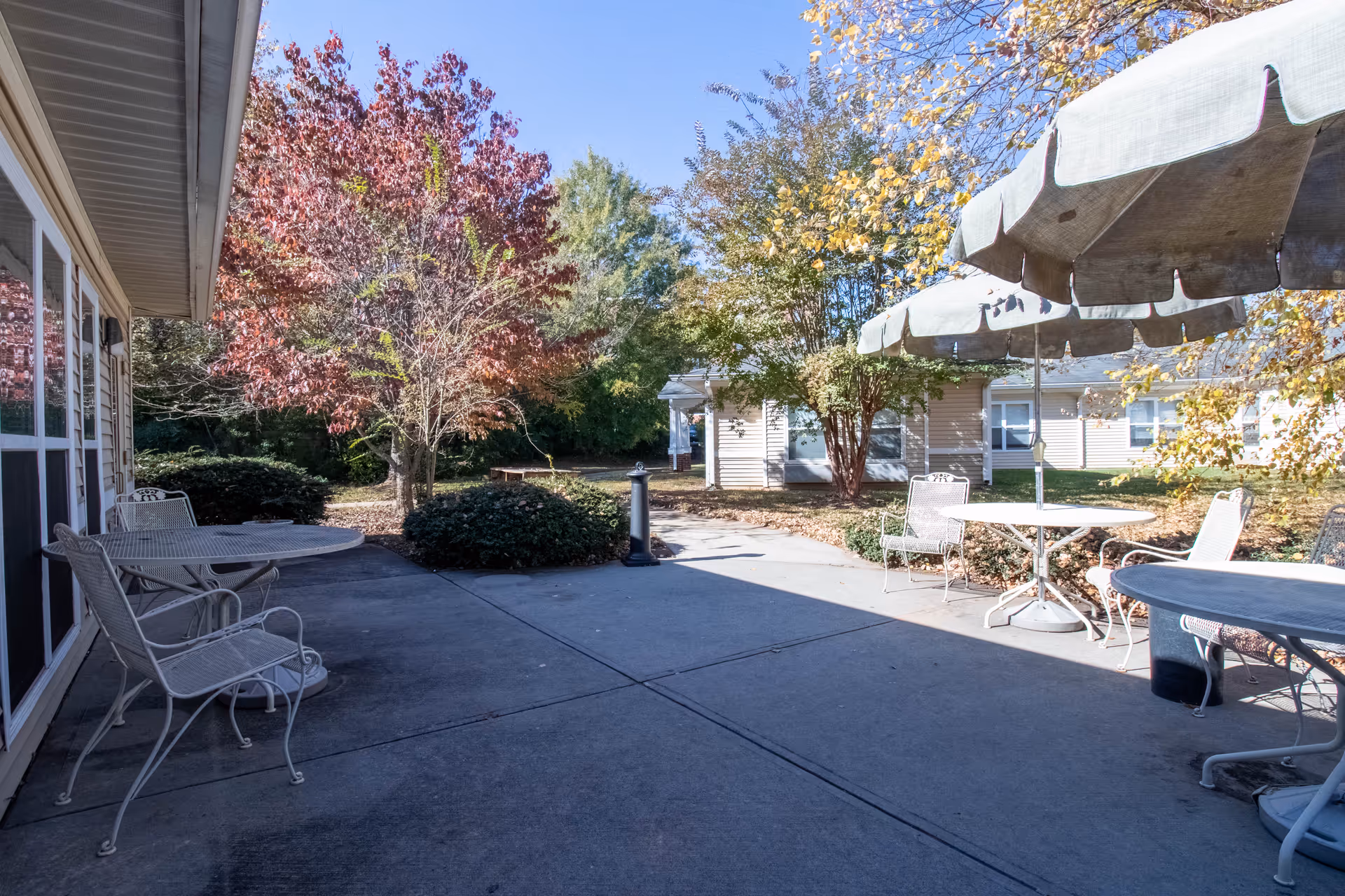 Outdoor patio area with metal tables and chairs, some tables shaded by large umbrellas. Surrounding the patio are trees with autumn-colored leaves and bushes, with a beige building visible in the background under a clear blue sky.