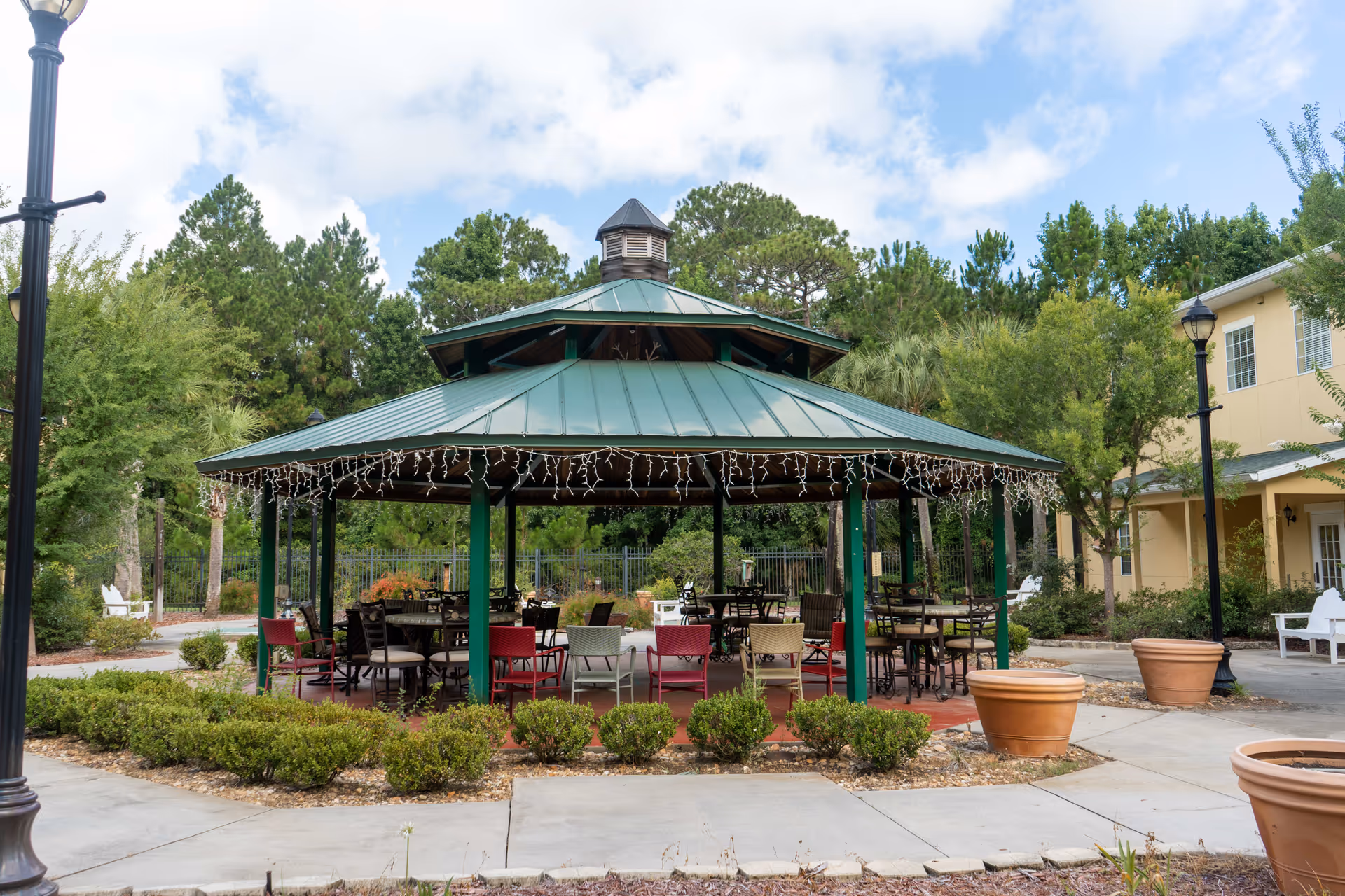 Outdoor gazebo with a green metal roof and string lights hanging from the edges, surrounded by chairs and tables. The gazebo is situated in a landscaped area with bushes, large potted plants, and trees in the background. There are lamp posts and a beige building visible on the right side under a partly cloudy sky.