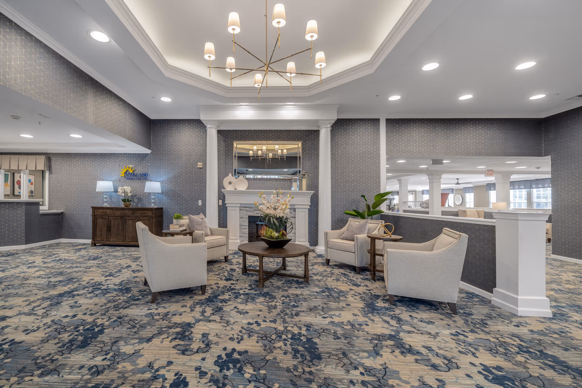 A spacious and elegant common area with a patterned blue and beige carpet, four beige armchairs arranged around a round wooden coffee table with a floral centerpiece, a white fireplace with decorative items and a large mirror above it, and a modern chandelier hanging from a recessed ceiling. The walls are covered in a subtle patterned wallpaper, and there is a wooden sideboard with two lamps and a plant on the left side.