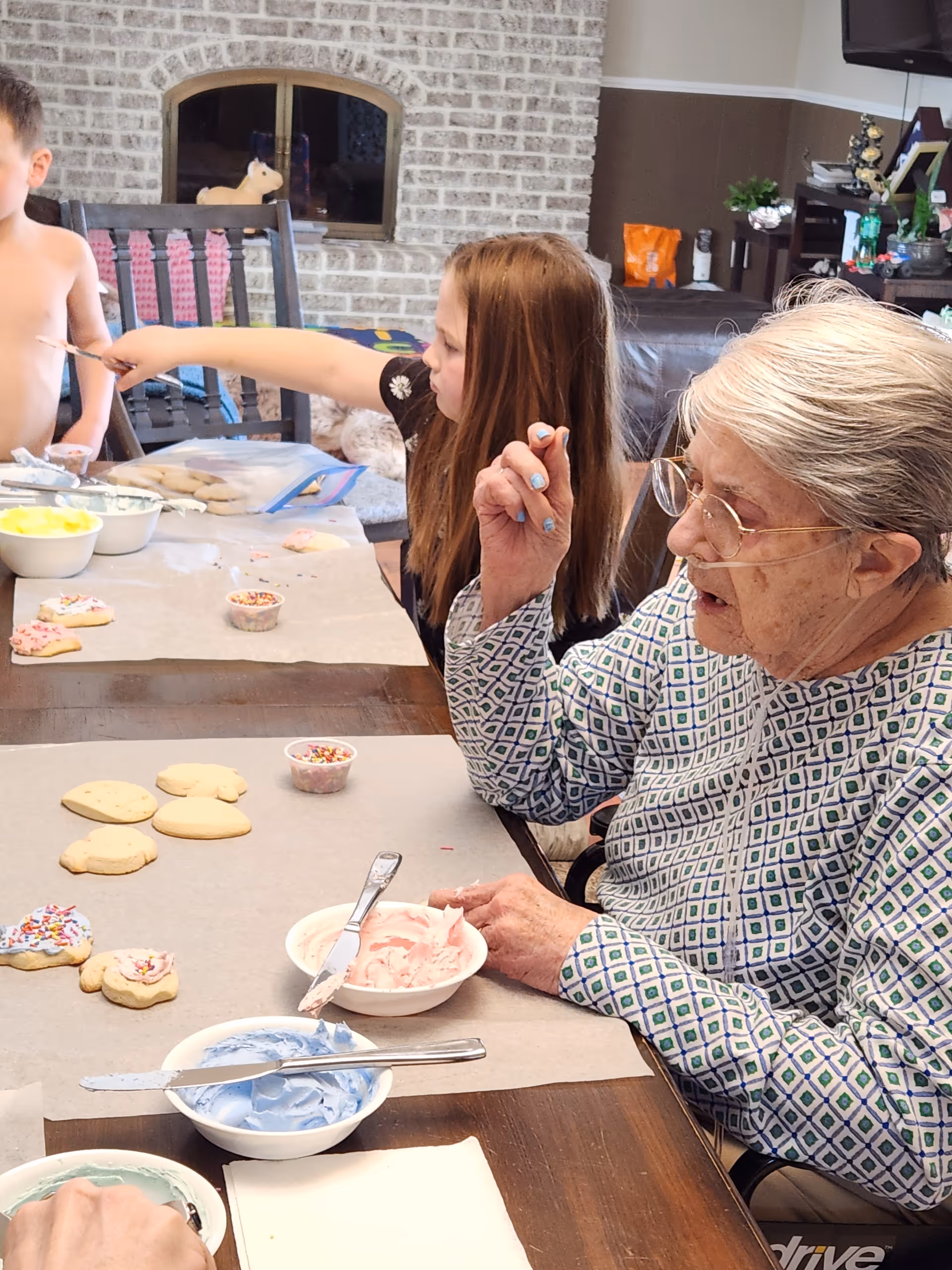 An elderly woman with glasses and a patterned shirt is sitting at a table decorating cookies with pink and blue icing. Two children are also at the table, one reaching across to spread icing. Bowls of colored icing and sprinkles are on the table. The background shows a brick fireplace and living room furniture.