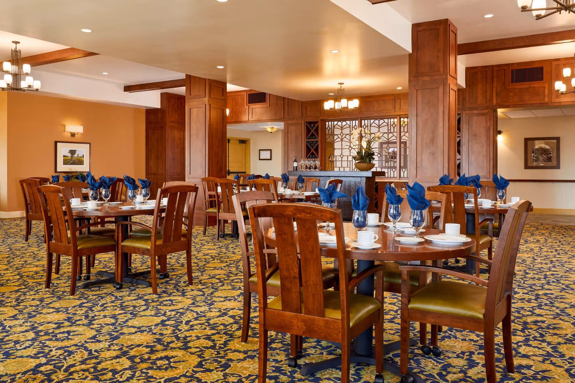 A dining room with multiple round wooden tables set with white plates, cups, and blue folded napkins in glasses. The room features patterned carpet in blue and yellow, wooden chairs with green cushions, warm lighting, and wood-paneled columns and walls.