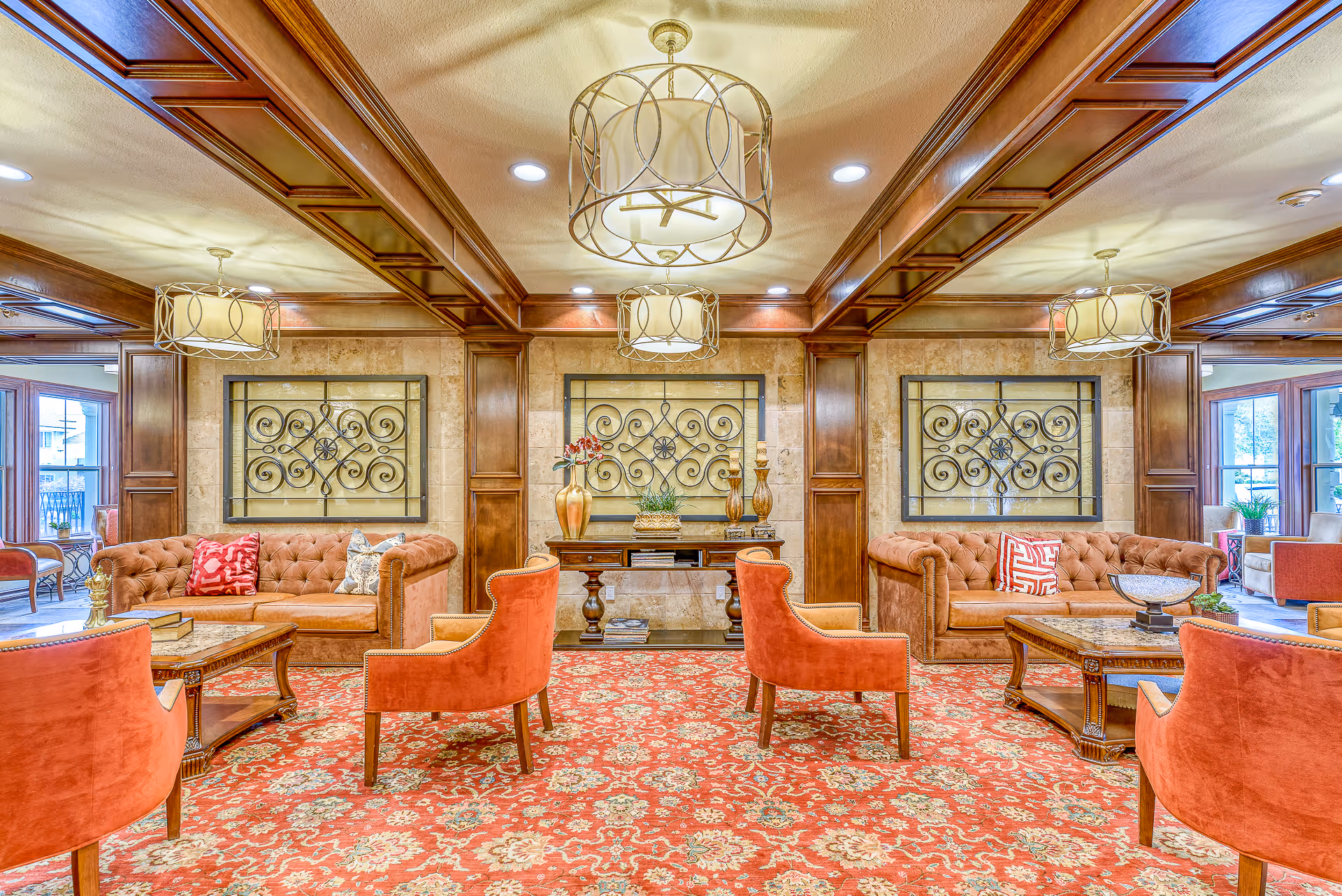 Elegant senior living lounge with tufted sofas, orange armchairs, decorative wall panels, chandeliers and patterned carpet.