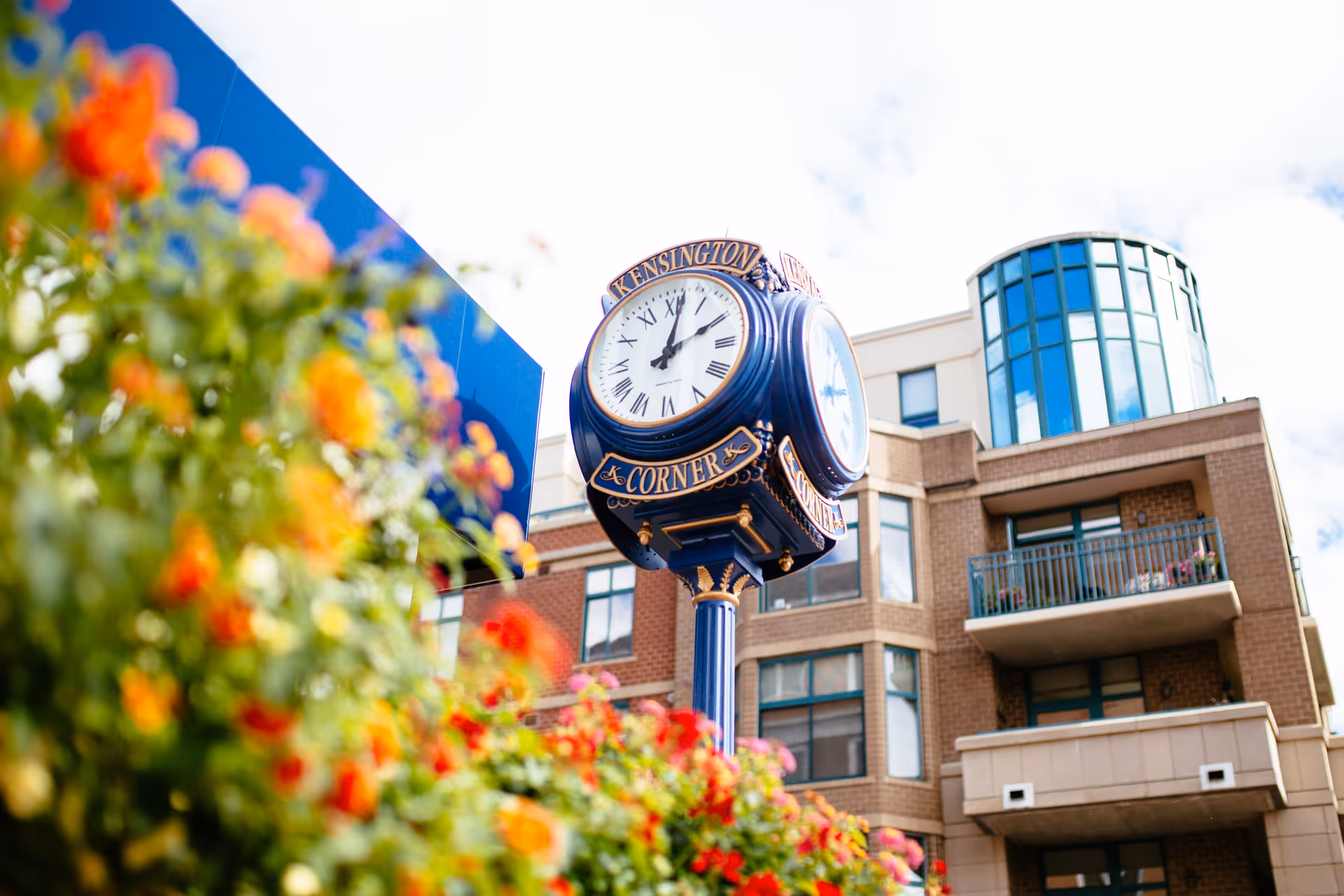 Decorative street clock reading 'Kensington Corner' in front of a multi-story brick building with balconies and colorful flowers in the foreground.