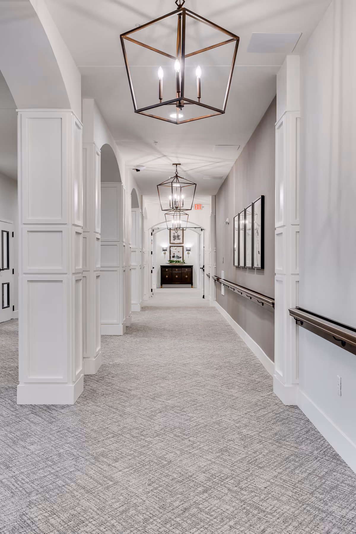 Bright carpeted hallway with white paneled columns, handrails, and hanging lantern-style lights leading to a console table at the far end.