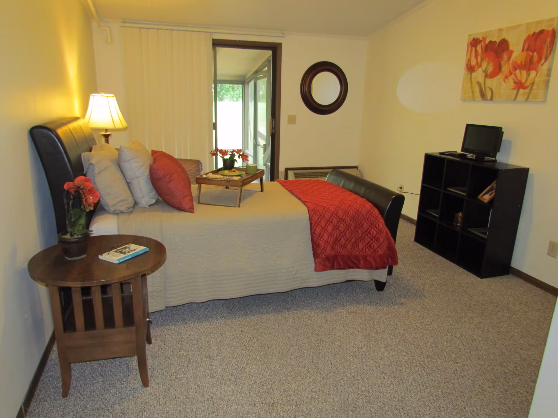 A cozy bedroom in an assisted living facility featuring a bed with beige bedding and red pillows and blanket. There is a wooden side table with a lamp and a potted plant, a small black shelving unit with a TV on top, a round mirror on the wall, and a floral painting. A sliding glass door leads to an enclosed porch or sunroom.