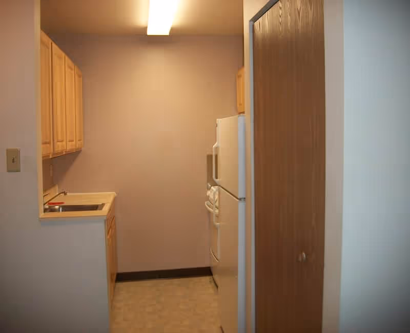 Small kitchen area with light wood cabinets, a sink, a refrigerator, and a stove. The walls are painted beige, and there is a wooden door on the right side.