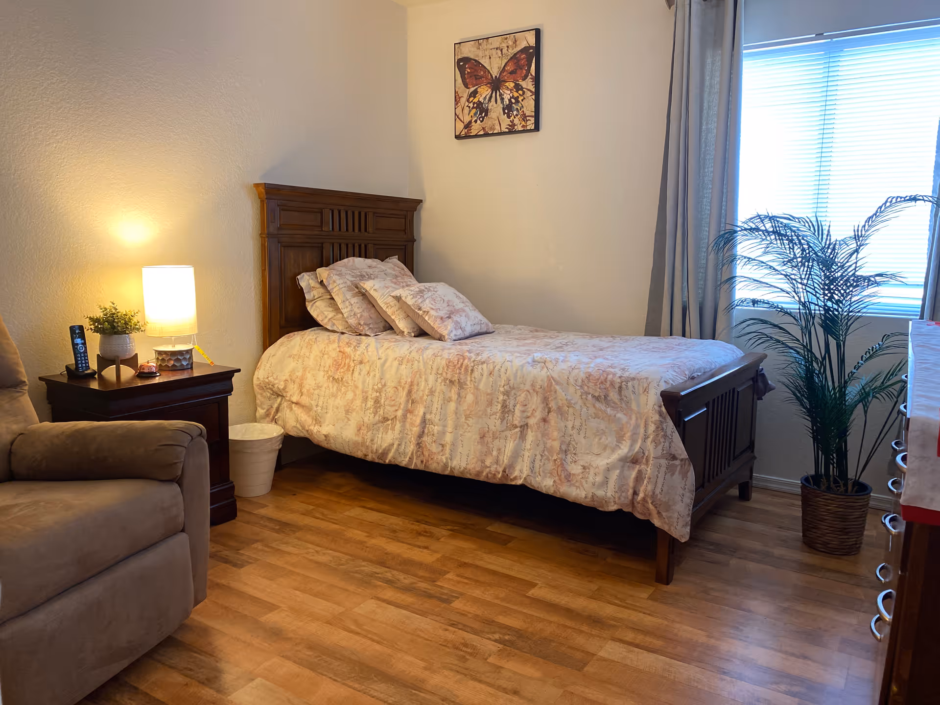 A cozy bedroom with a wooden bed frame and a bed made with floral patterned bedding and multiple pillows. Next to the bed is a wooden nightstand with a lit lamp, a small plant, and a remote control. There is a beige armchair partially visible on the left side. On the wall above the bed, there is a framed butterfly artwork. A window with blinds and gray curtains is on the right side, next to a potted plant on the wooden floor.