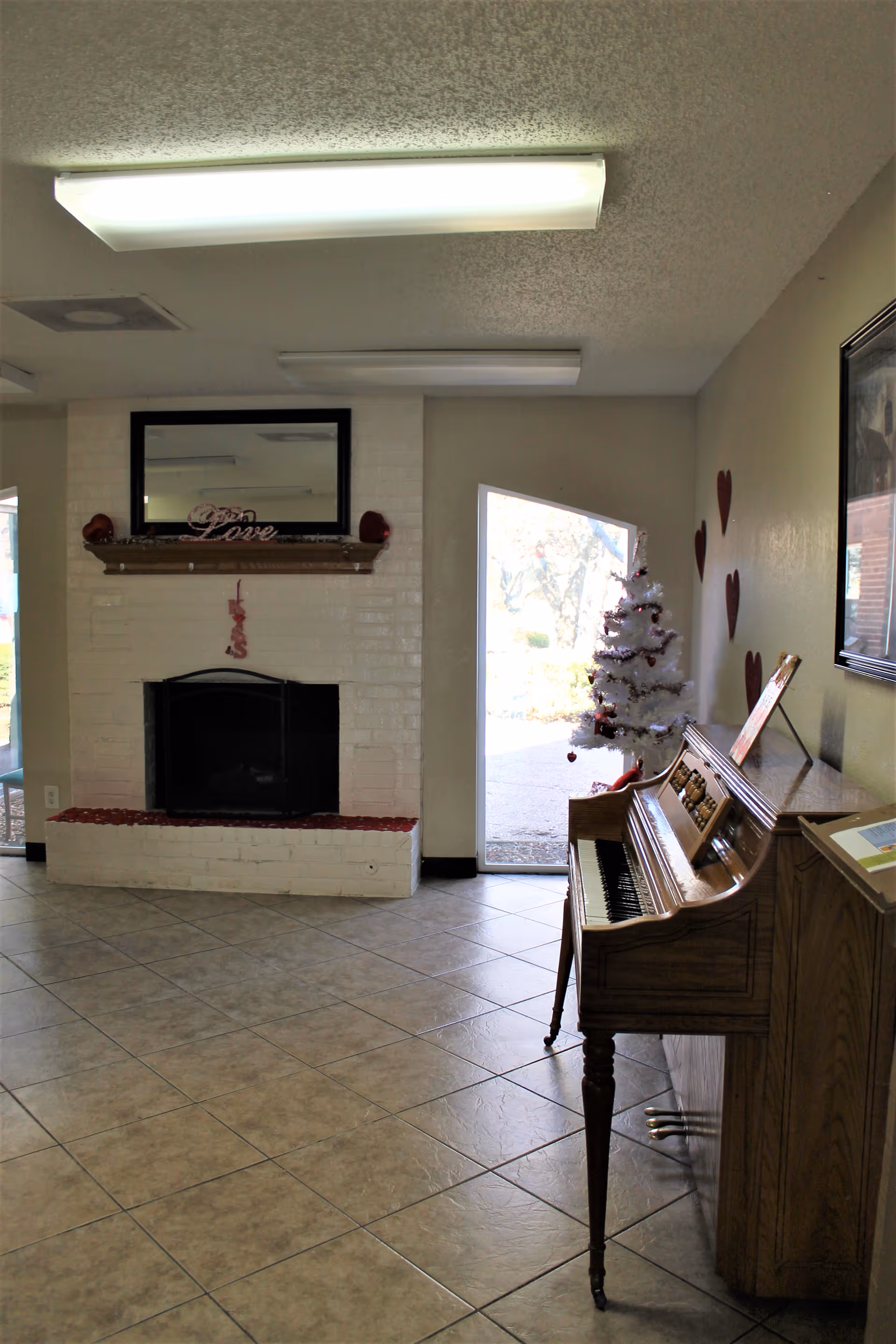 Interior room with a white brick fireplace decorated with a 'Love' sign and small red ornaments. To the right, there is a small white Christmas tree with red decorations and a wooden piano with sheet music on top. The floor is tiled, and there are heart decorations on the wall. A door next to the Christmas tree leads outside, letting in natural light.