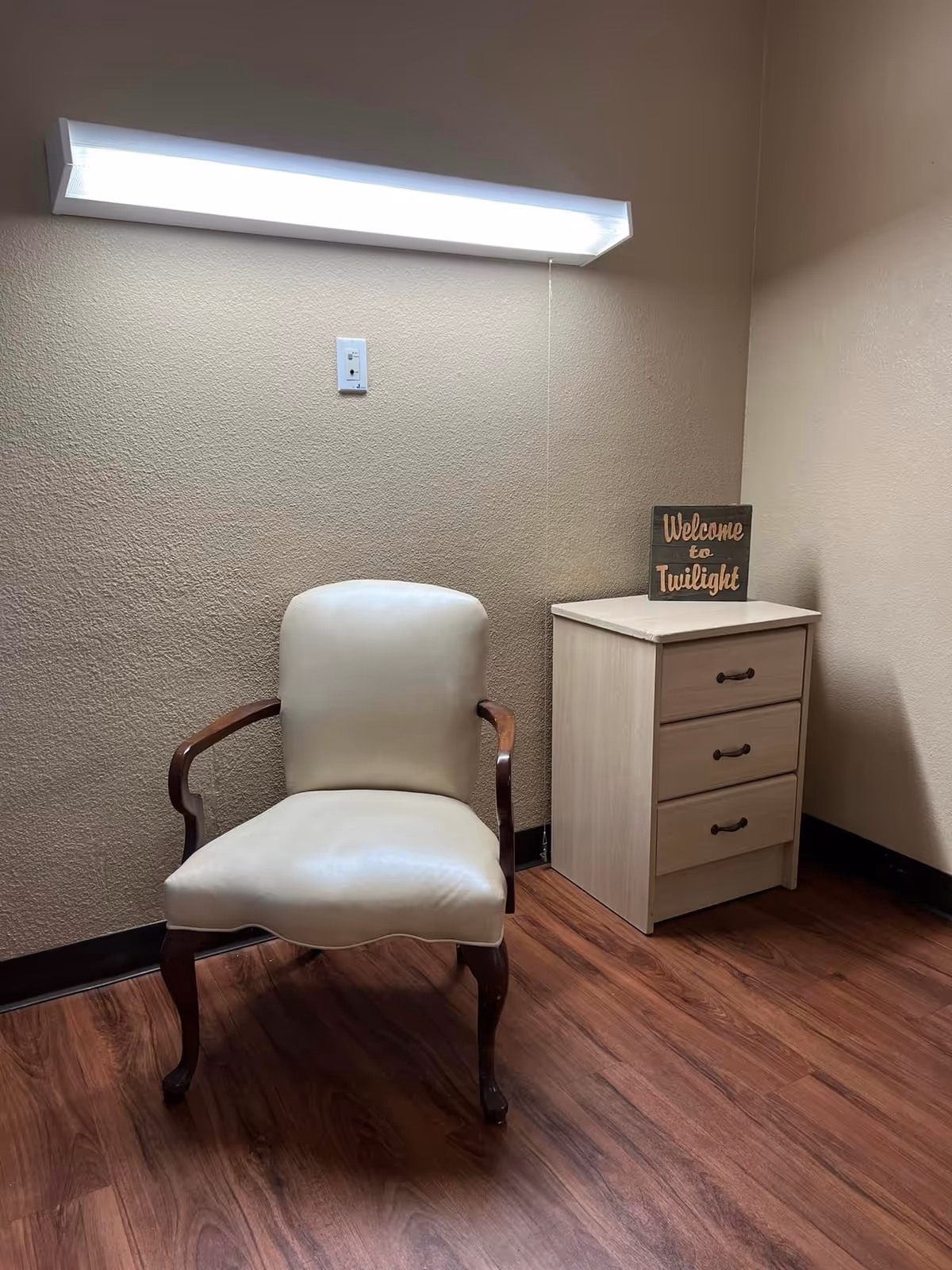 A small corner of a room with a beige upholstered wooden armchair on a wood floor next to a light-colored wooden three-drawer nightstand. On top of the nightstand is a small sign that reads 'Welcome to Twilight'. A long fluorescent light fixture is mounted on the beige textured wall above the chair and nightstand.