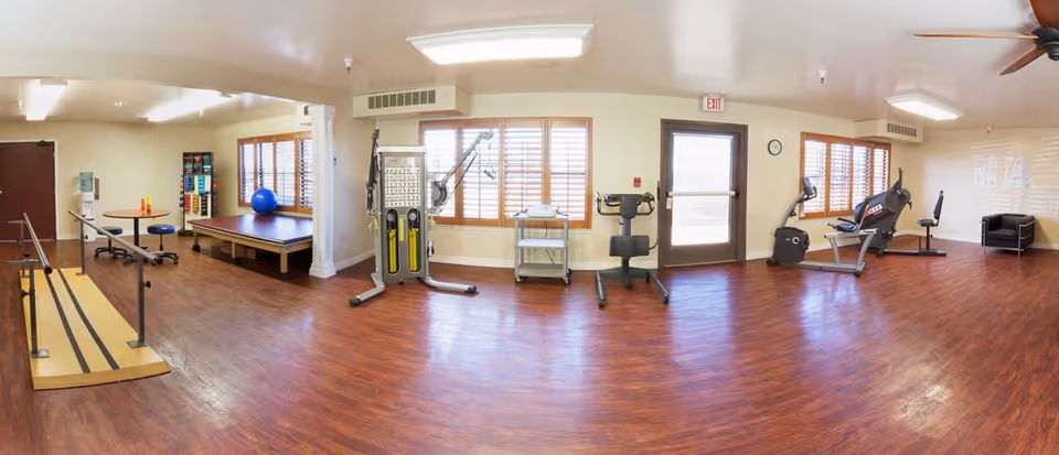 A spacious rehabilitation or physical therapy room with wooden flooring, featuring parallel bars for walking exercises, a padded therapy table with a blue exercise ball, various exercise and therapy equipment, two stationary exercise bikes, a small table with chairs, and a black armchair near the windows with wooden blinds. The room is well-lit with ceiling lights and has an exit door.