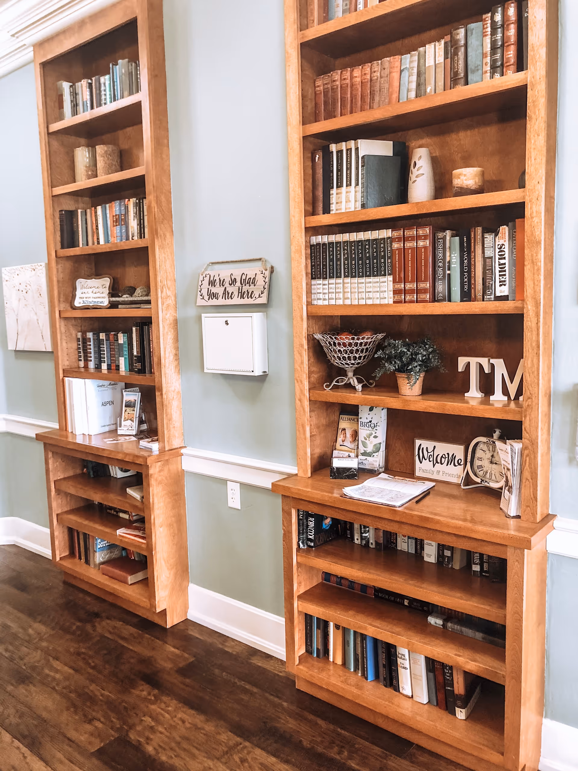 Interior view of a hallway with wooden bookshelves filled with books, decorative items, and welcome signs. The walls are painted light green with white trim, and the floor is dark wood.