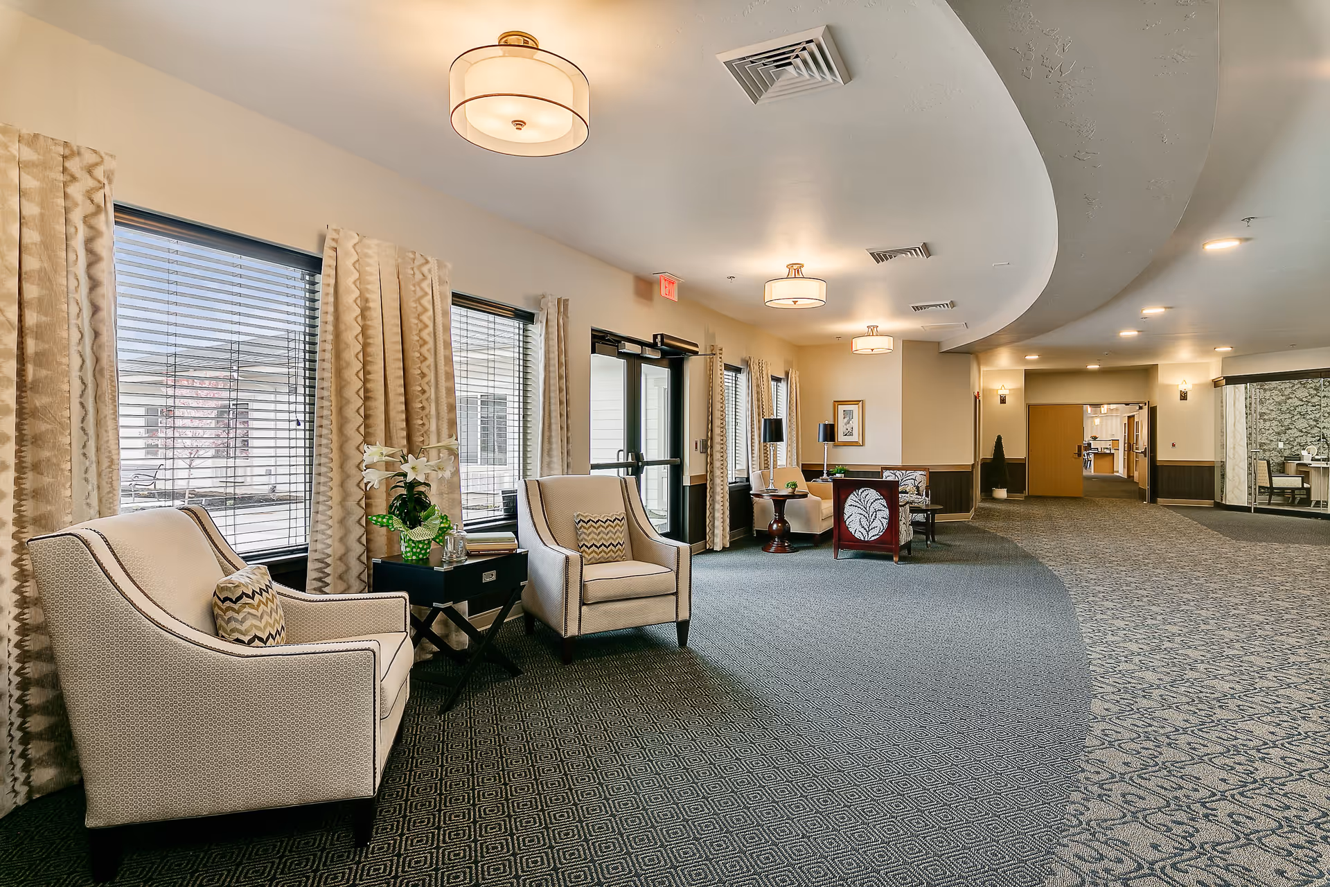 A spacious, well-lit senior living facility common area with patterned carpet, beige walls, and large windows with curtains. The seating area includes several upholstered armchairs with decorative pillows, small tables with lamps and a flower arrangement. Ceiling lights and air vents are visible, and a hallway leads to other parts of the facility.