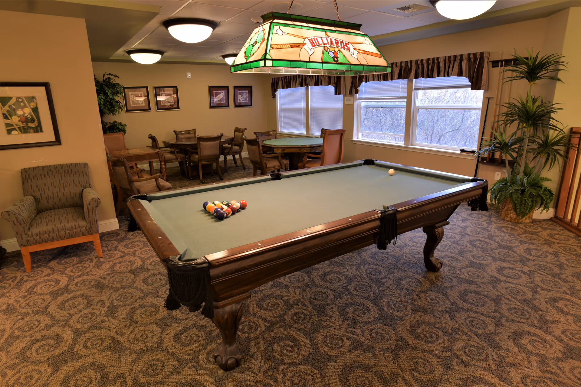 A recreational room with a pool table in the center, set up for a game with balls racked and a cue ball placed. Above the pool table hangs a stained glass light fixture with the word 'Billiards'. In the background, there are several tables and chairs arranged for seating, framed pictures on the wall, large windows with blinds, and some potted plants. The room has patterned carpet flooring and warm lighting.
