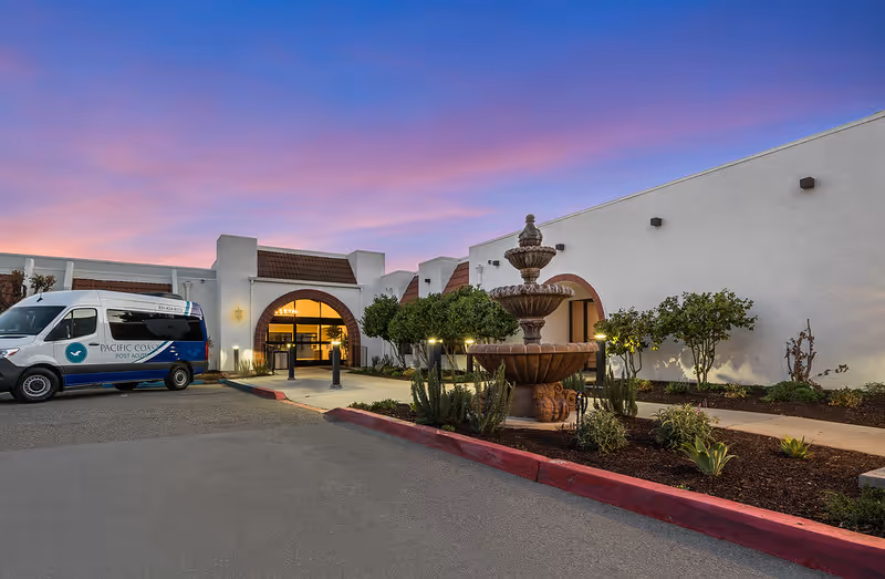 Exterior view of Pacific Coast Post Acute facility at dusk with a multi-tiered fountain in the landscaped area near the entrance and a facility shuttle van parked on the left side.