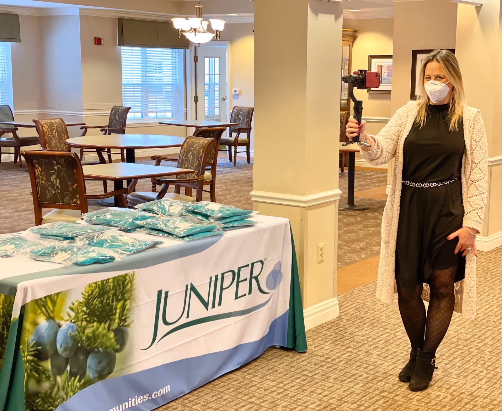 A masked woman holding a camera gimbal stands beside a table with a Juniper-branded tablecloth and packaged items in a dining/activity room.