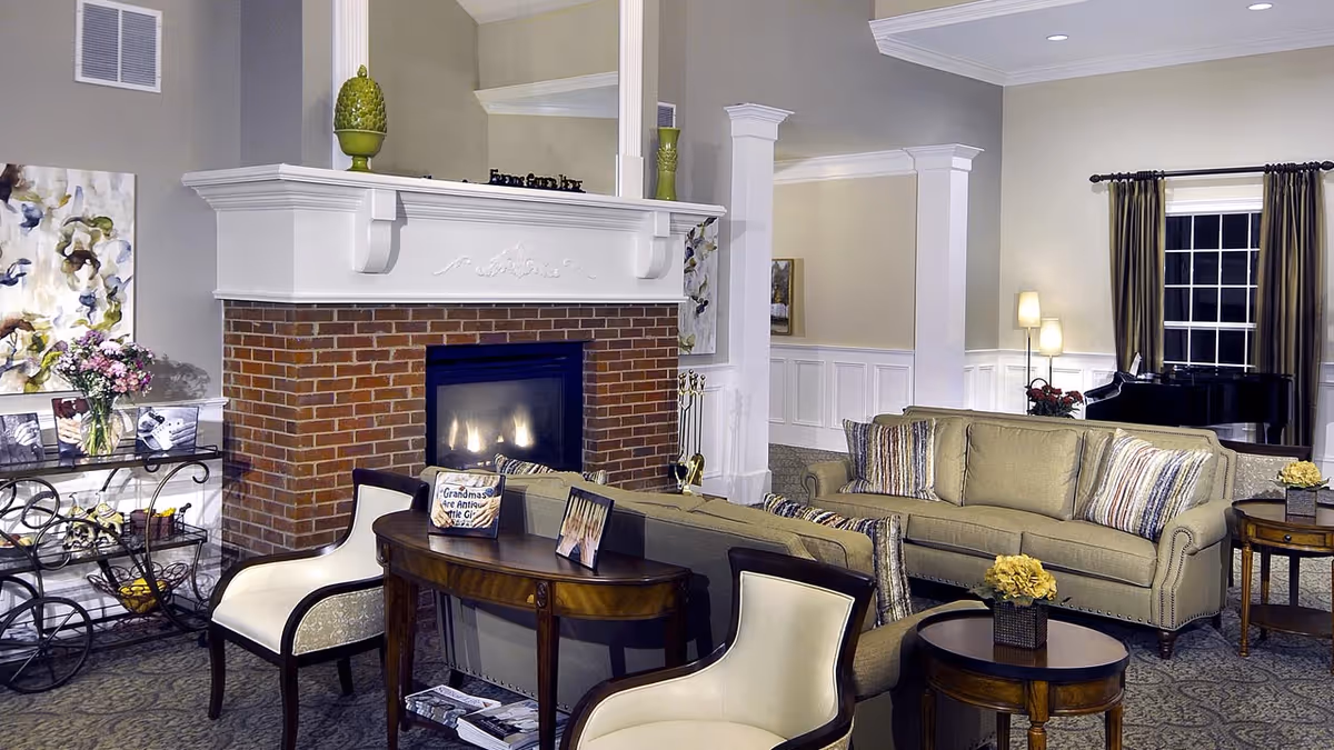 A cozy senior living room featuring a brick fireplace with a white mantel, two beige sofas with striped cushions, two cream-colored armchairs, a wooden console table with framed photos, a side table with a small flower arrangement, a floor lamp, and a black grand piano near a window with brown curtains.