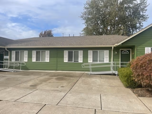 Single-story green building with white window shutters and two wheelchair ramps leading to separate entrances, surrounded by a concrete driveway and some bushes, under a partly cloudy sky.