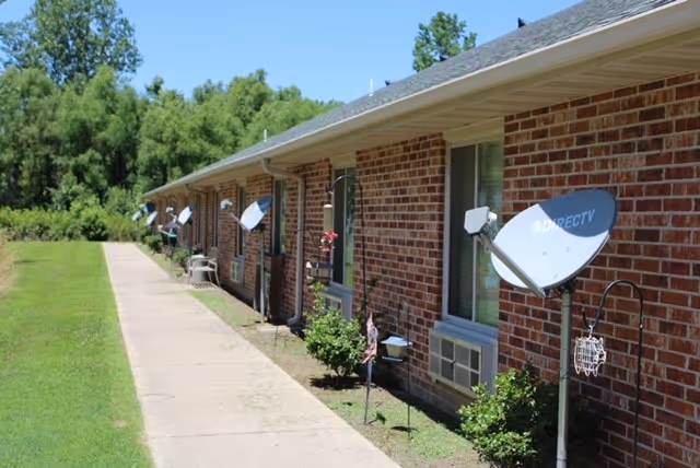 Brick single-story exterior of a care facility with a sidewalk, row of windows, satellite dishes, and a grassy lawn.