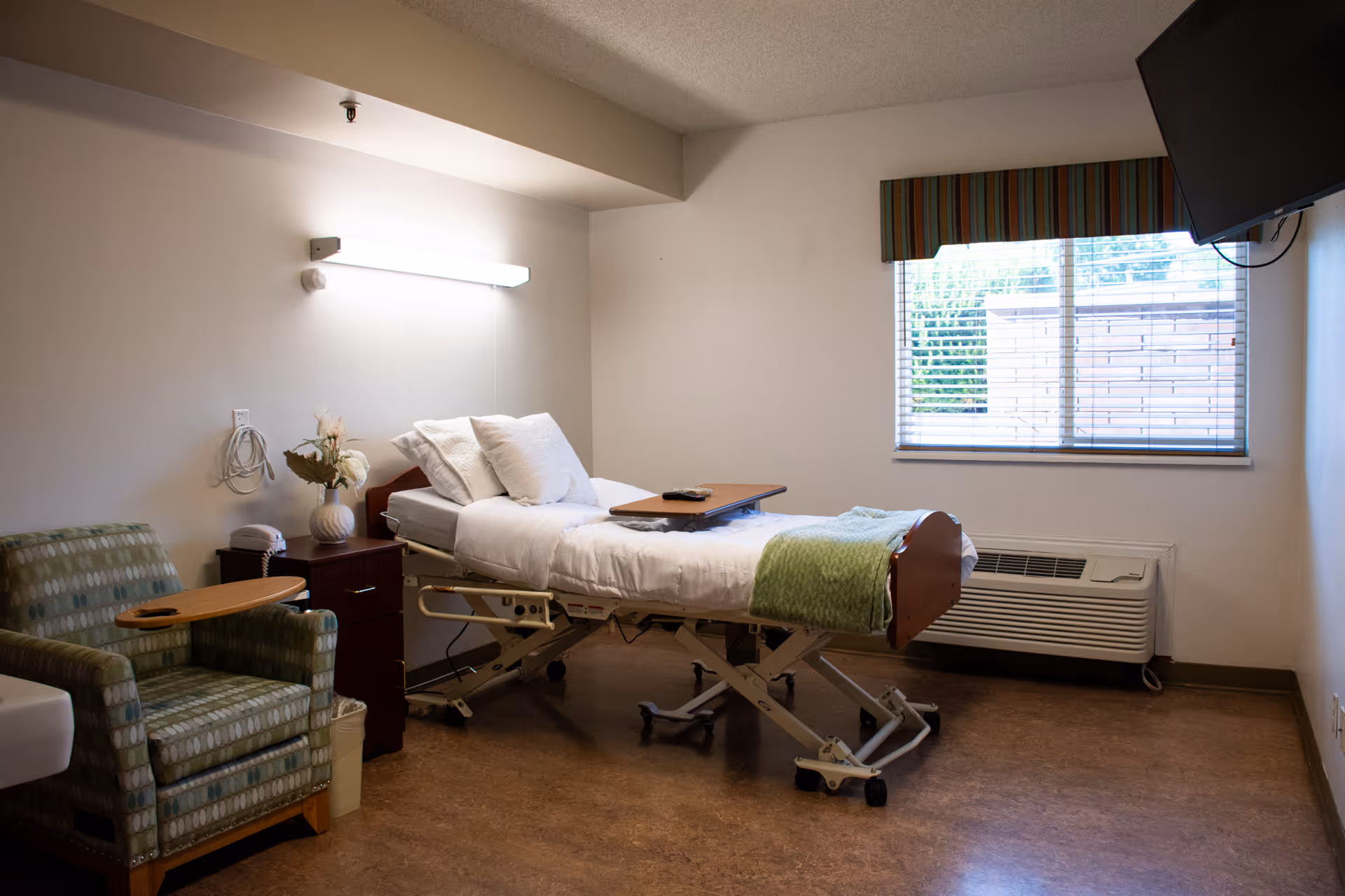 Sparsely furnished patient bedroom with a hospital-style bed, bedside table, armchair and a window with blinds.