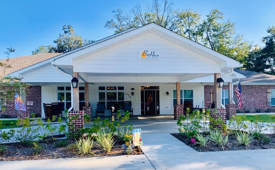 Front exterior view of BeeHive Homes Assisted Living facility with a covered entrance supported by wooden posts and brick bases, surrounded by landscaped plants and greenery under a clear blue sky.