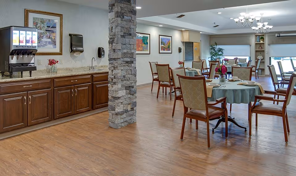 A dining room with wooden floors and several round tables covered with tablecloths and set with plates and cups. Chairs with wooden frames and cushioned seats surround the tables. On the left side, there is a counter with a beverage dispenser, a sink, and cabinets underneath. The room has framed artwork on the walls, a stone pillar, and large windows with blinds letting in natural light.