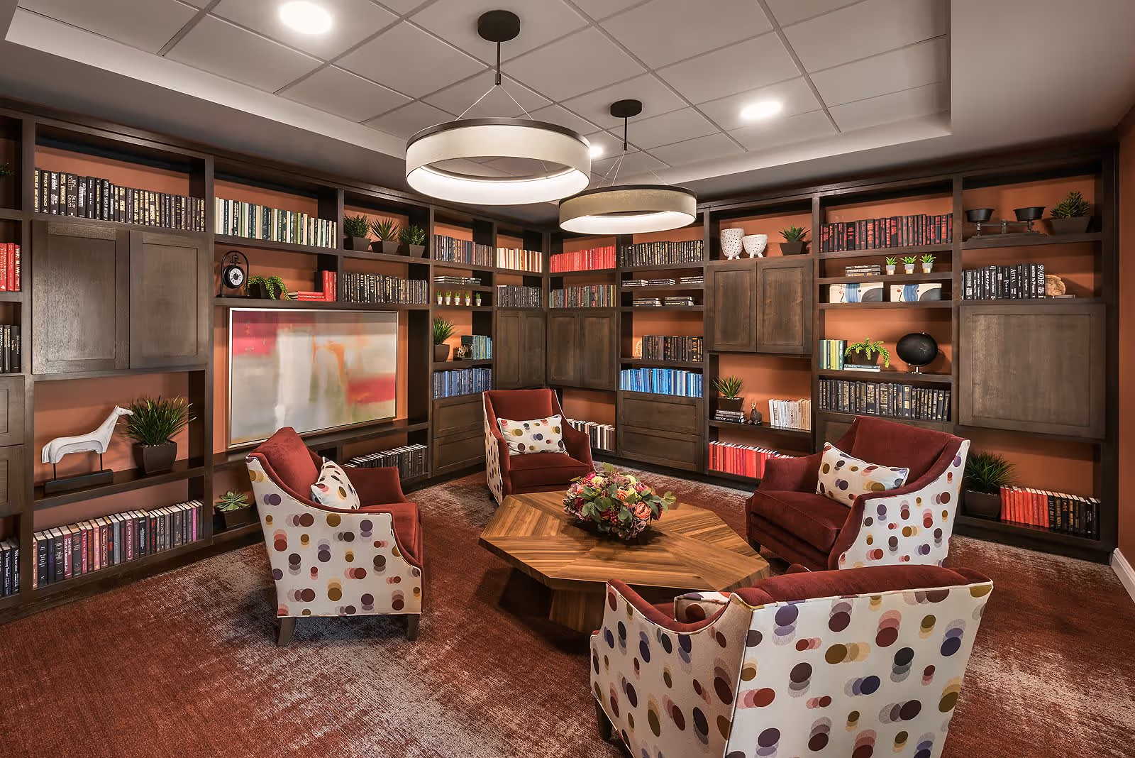 A cozy library or reading room with built-in dark wood bookshelves filled with books and decorative plants. Four upholstered armchairs with red cushions and patterned backs surround a wooden hexagonal coffee table with a floral centerpiece. The room has warm lighting from two large circular ceiling fixtures and a carpeted floor in shades of red and brown.