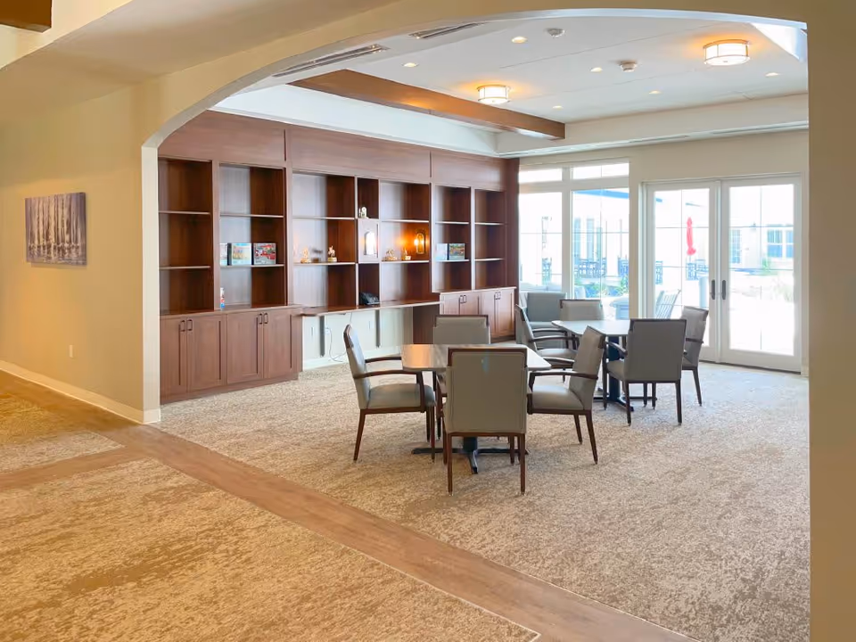 A bright and spacious common area in a senior living facility featuring a carpeted floor, wooden built-in shelves with decorative items, several tables with chairs, and large glass doors leading to an outdoor patio area.