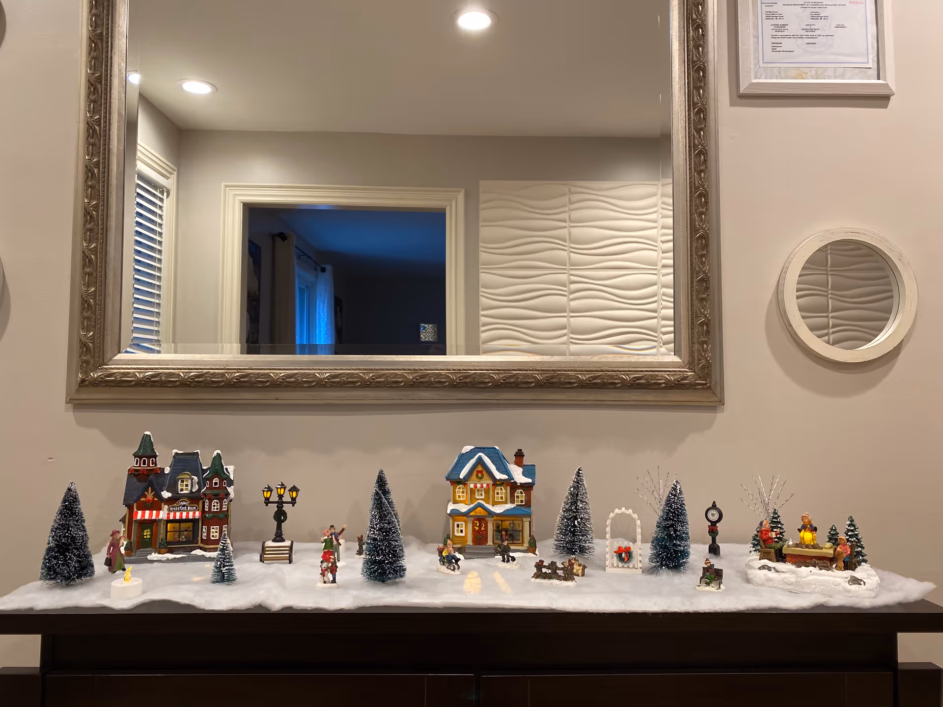 A decorative winter village scene displayed on a dark wooden surface with artificial snow, miniature houses, trees, street lamps, and small figurines. The scene is set against a beige wall with a large ornate mirror reflecting part of a room and a small round mirror to the right.