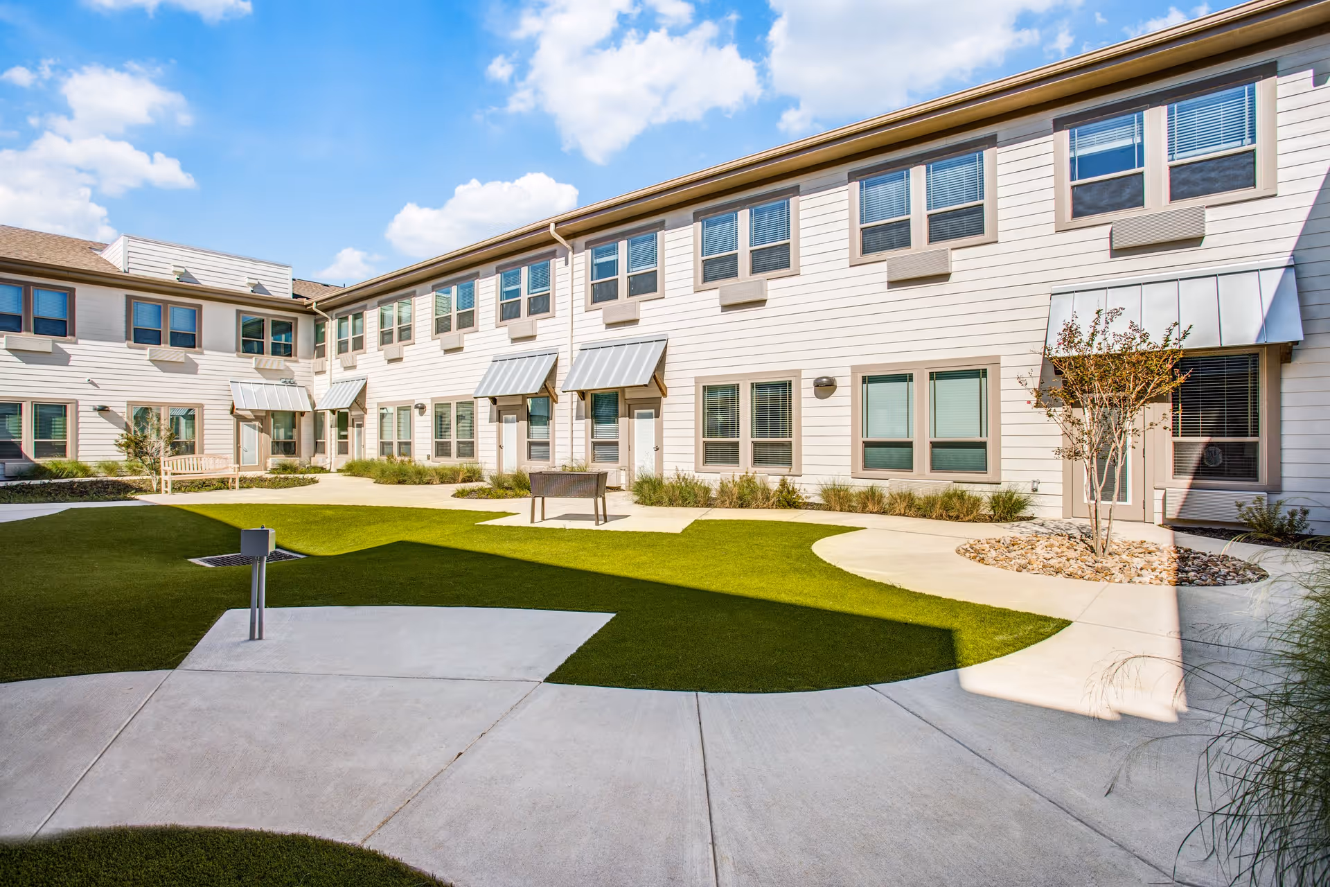 Outdoor courtyard area of a senior living facility with a well-maintained lawn, concrete walkways, small trees, and a two-story building with multiple windows and awnings under a blue sky with scattered clouds.