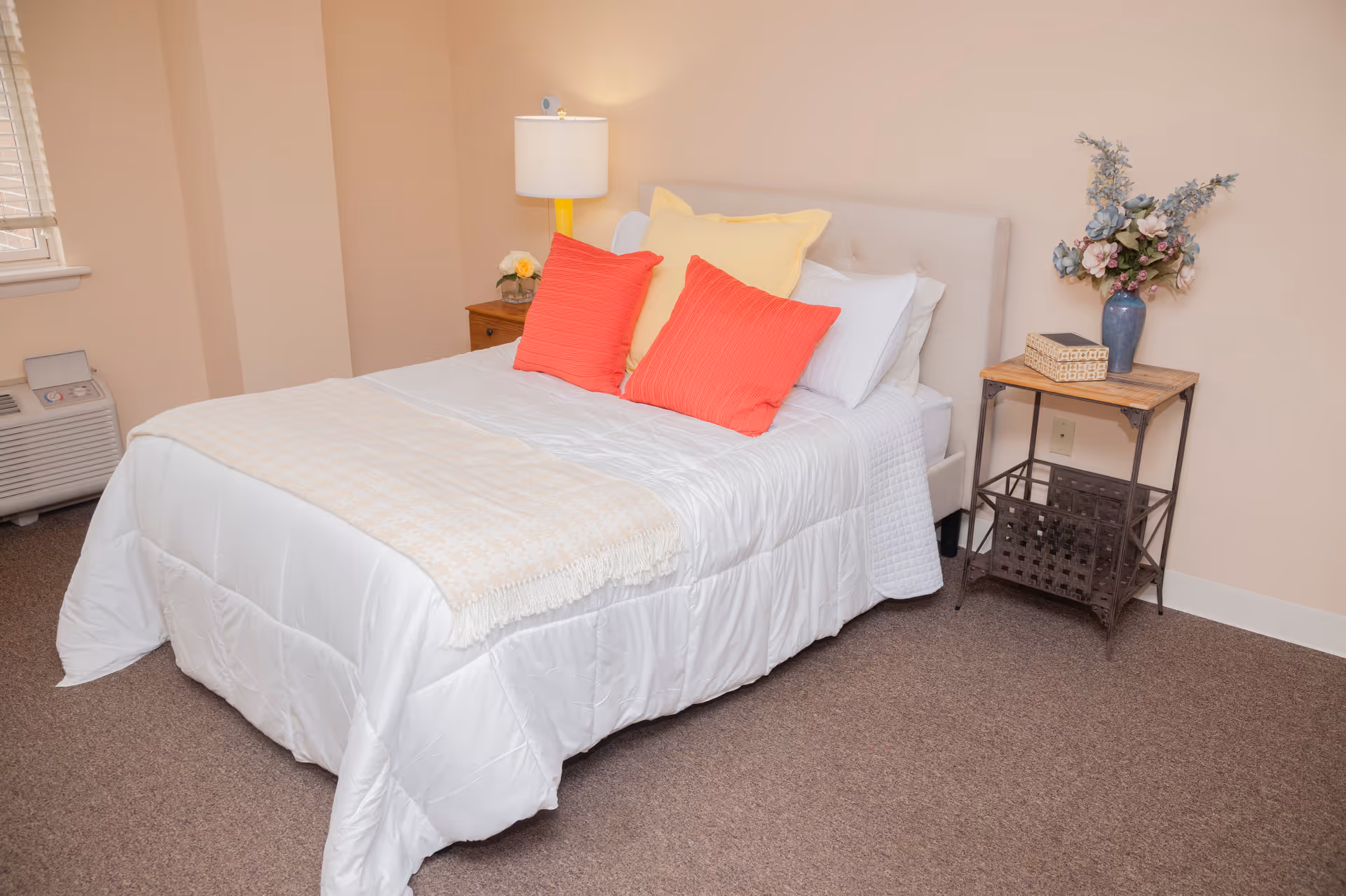 A neatly made bed with white bedding, two red pillows, and one yellow pillow in a softly lit bedroom. There is a wooden nightstand with a lamp and a small flower arrangement on one side, and a small table with a vase of flowers and a basket on the other side. The room has beige walls and carpeted floor.
