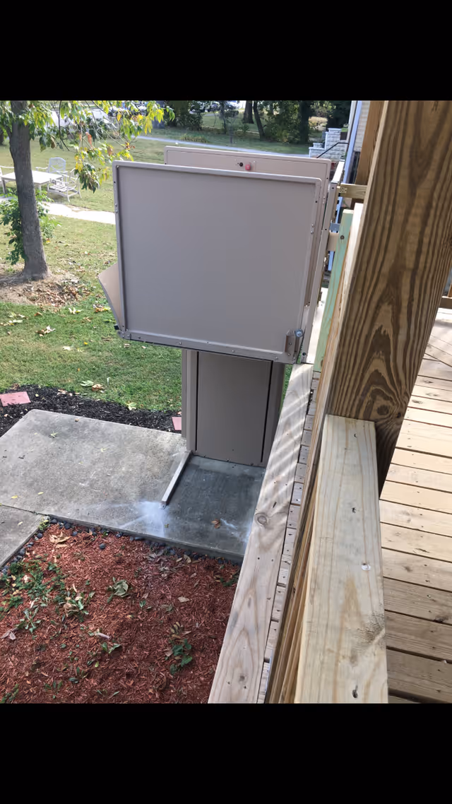 An outdoor wheelchair/platform lift attached to a wooden deck overlooking a concrete walkway and lawn.